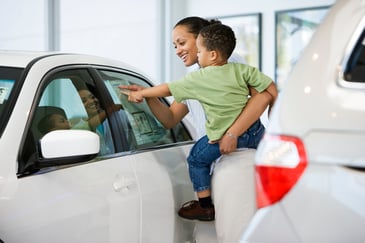 Woman holding a young child while they look at a car window sticker together at a dealership, smiling and pointing at the details.