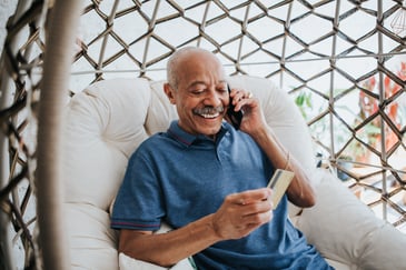Smiling older man sitting on a cushioned patio chair, holding a credit card and talking on his phone, with a relaxed and cheerful expression.