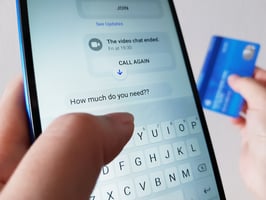 Close-up of a person holding a smartphone and a blue credit card. The phone screen shows a messaging app with a video call ended and a message saying, “How much do you need??”, suggesting a potential romance scam.