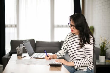 A woman with a pen, paper, and a laptop works on her finances.