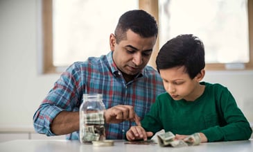 Father and son counting their savings together at a table, with a jar full of cash and coins in front of them.