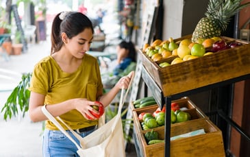 Young woman placing fresh produce into a reusable tote bag at an outdoor fruit and vegetable stand, promoting eco-friendly and sustainable shopping habits.