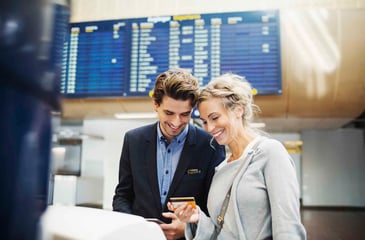 Smiling couple at an airport checking travel details on a phone and credit card, with a departures board in the background.