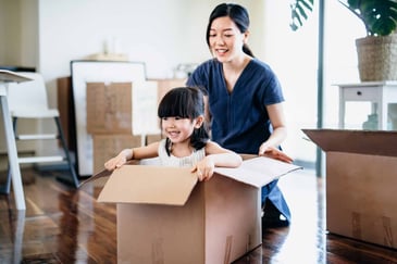 Smiling mother and daughter surrounded by moving boxes, with the child playing inside one, suggesting a recent home move or new beginning.