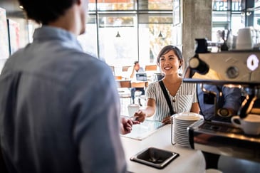 Barista smiling as she accepts a card payment from a customer at a coffee shop counter, with a coffee machine and takeaway cup nearby.