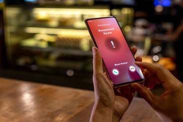 Person holding a smartphone displaying an incoming call screen labeled 'Suspected Spam,' with options to answer or block the call.