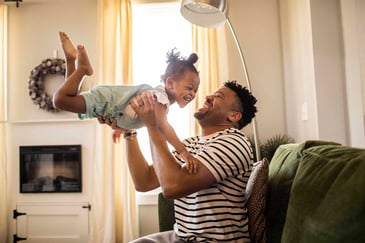 Father joyfully lifting and playing with his young daughter in a cozy living room, both smiling and enjoying quality time together.