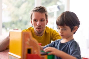 Father watching his young son play with a toy house, engaging in a focused and educational moment at home.
