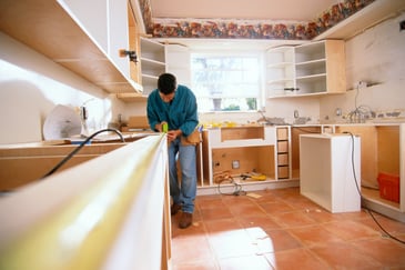 Man measuring and installing cabinetry during a kitchen renovation, surrounded by tools and unfinished surfaces.