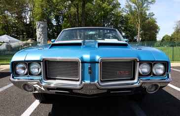 Front view of a classic blue Oldsmobile 442 muscle car parked at an outdoor event, with trees and vendor tents in the background.