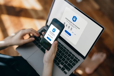 Person using a smartphone and laptop for two-factor authentication, entering a six-digit verification code displayed on the phone into a login screen on the laptop.