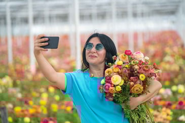 Woman taking a selfie while holding a colorful bouquet of flowers in a vibrant flower field, wearing sunglasses and a blue shirt.
