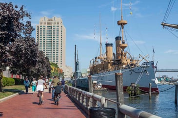 People biking and walking along the Philadelphia waterfront near historic ships, with the city skyline in the background.