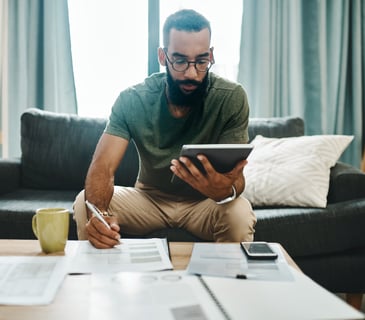 Man reviewing financial documents at home with a tablet in hand, surrounded by paperwork and a coffee mug on the table.