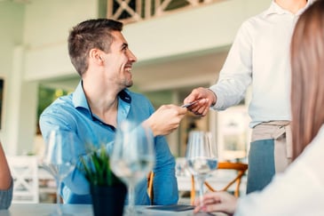 A smiling young man hands a waiter his credit card at a restaurant.